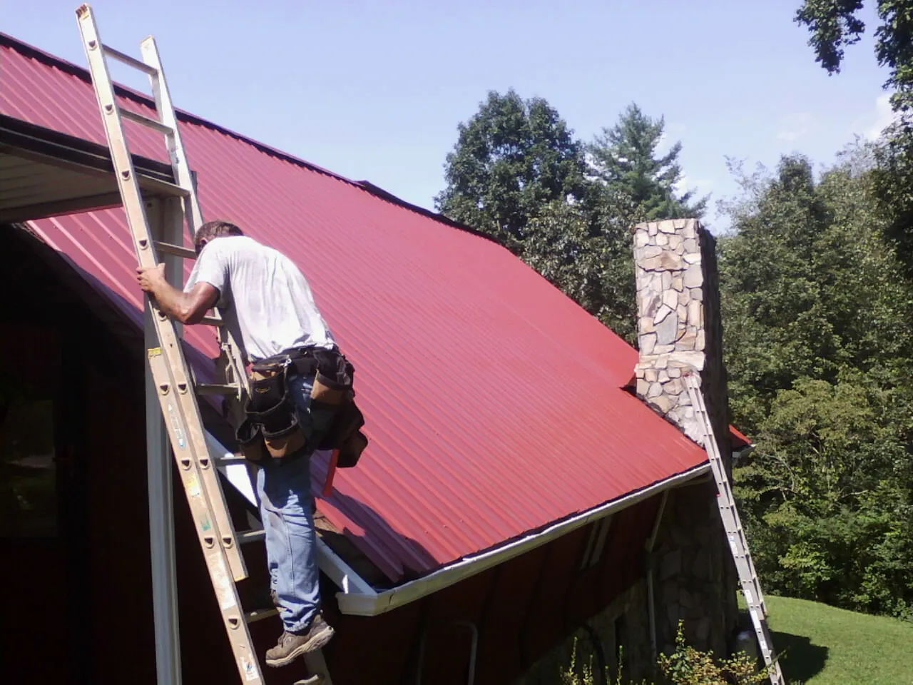 Roofer on a ladder beside a red metal roof and stone chimney — Soffit & Fascia Repair by Roofing Brothers in Chester Springs